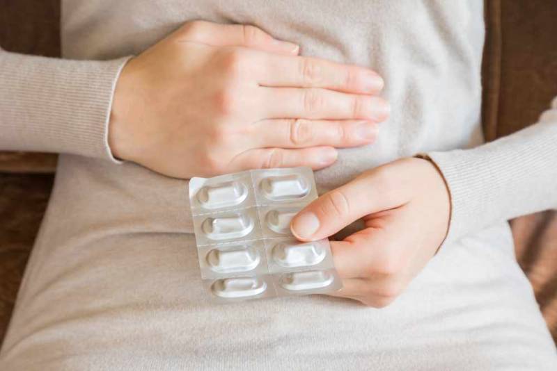 A woman holding her stomach with one hand while holding a strip of medication in the other, indicating relief or treatment for digestive discomfort