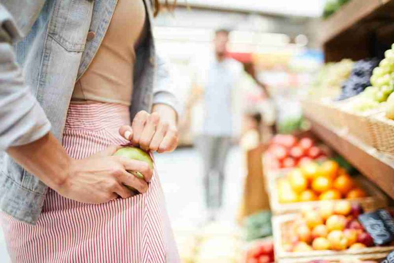 Woman secretly taking fruits from a supermarket shelf, illustrating compulsive stealing behavior caused by Kleptomania, a psychological impulse control disorder