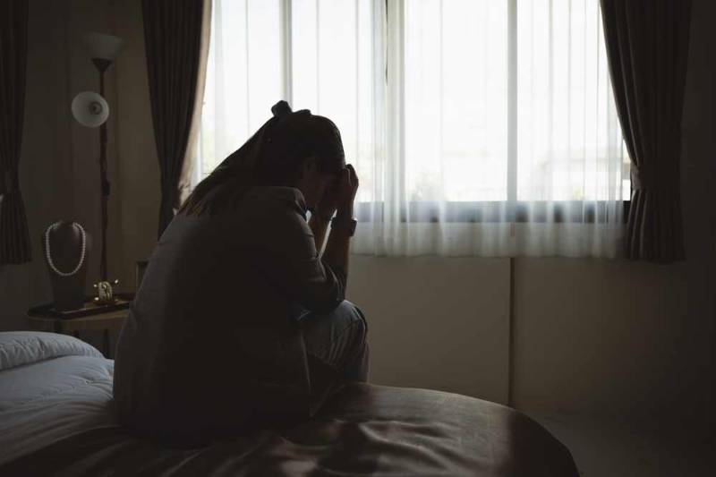 A woman sitting alone on a bed in a dark room, appearing withdrawn and overwhelmed, illustrating depression and emotional distress