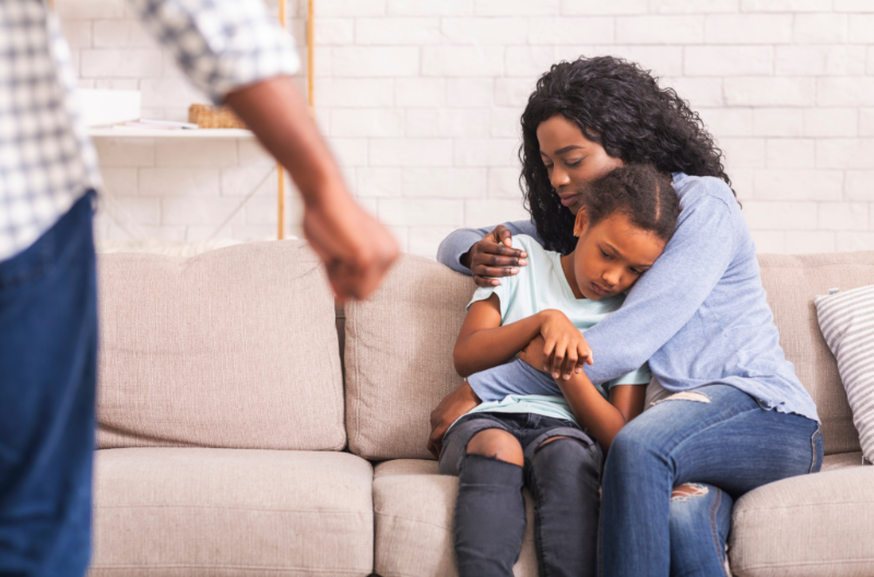 A distressed mother hugs her young daughter tightly while the angry husband shouts in the background, illustrating domestic violence at home